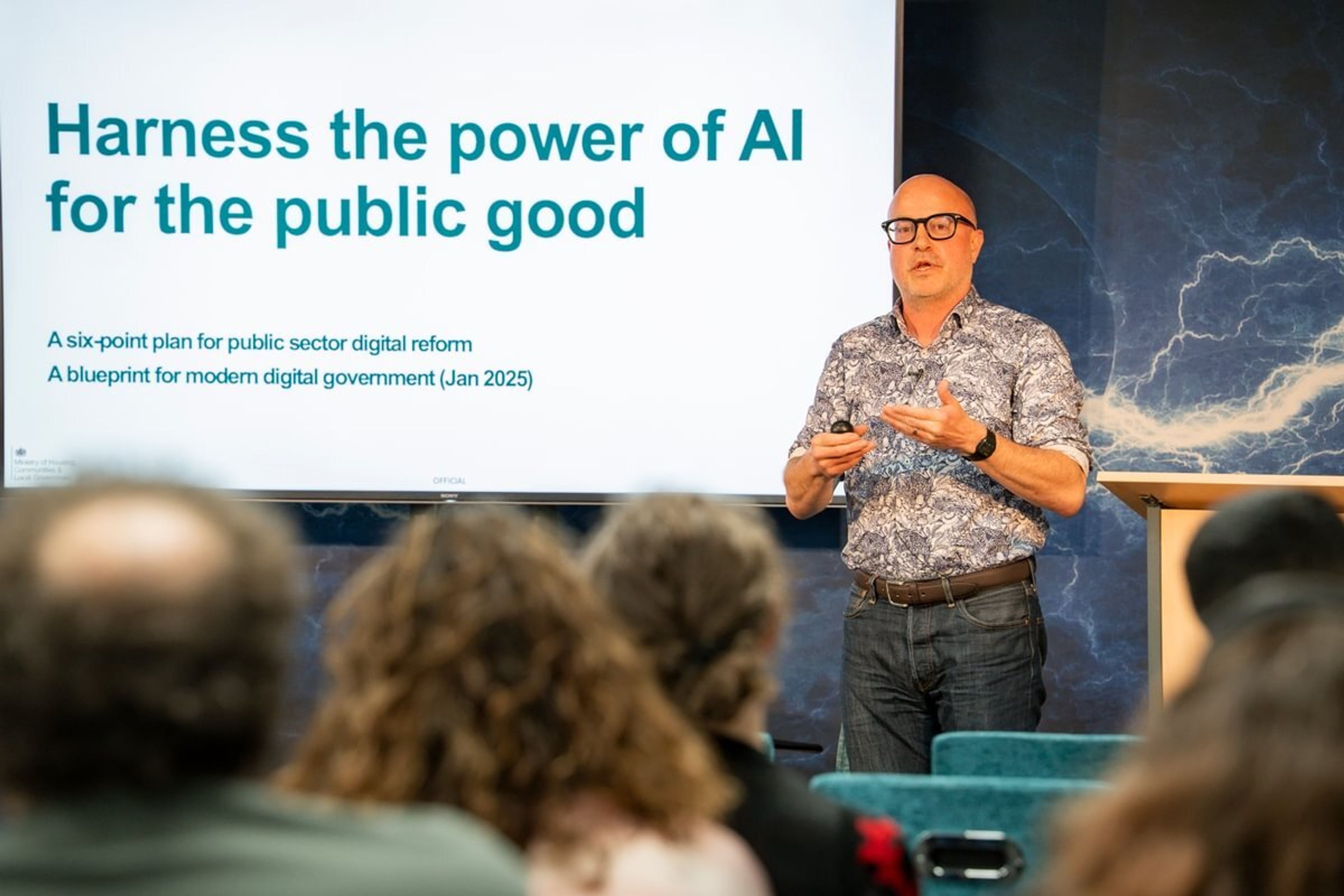 Tom Smith delivering his data bites presentation next to a screen that reads "harness the power of AI for the public good".