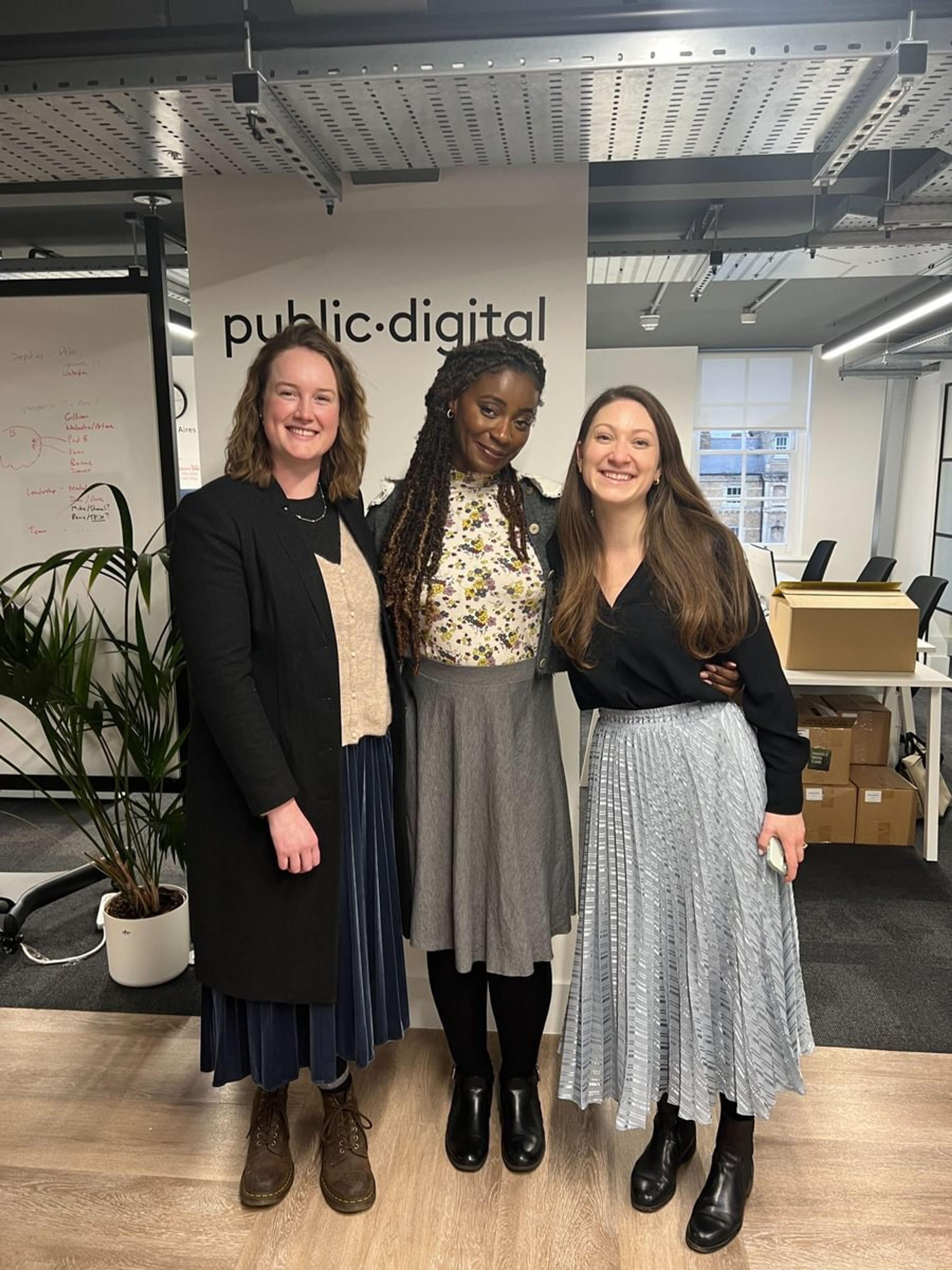 Three women pose to camera in front of Public Digital's logo