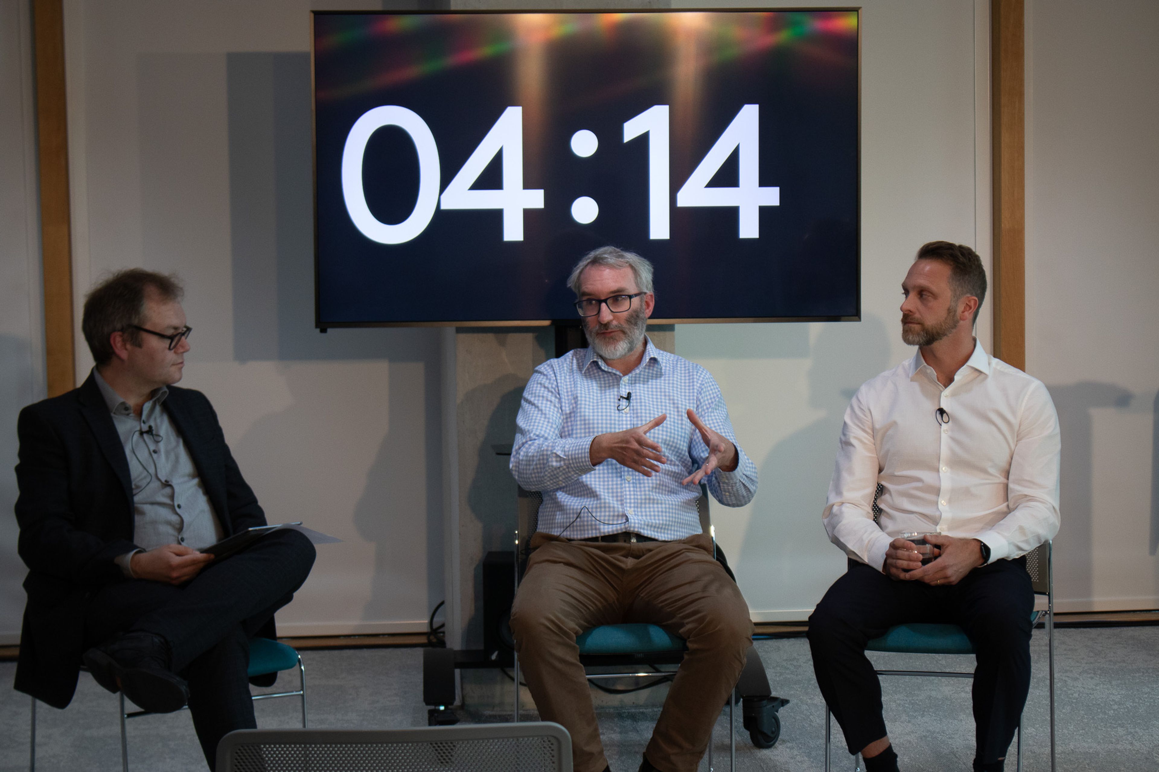Three men seating in chairs and discussing, with a screen showing time counting down