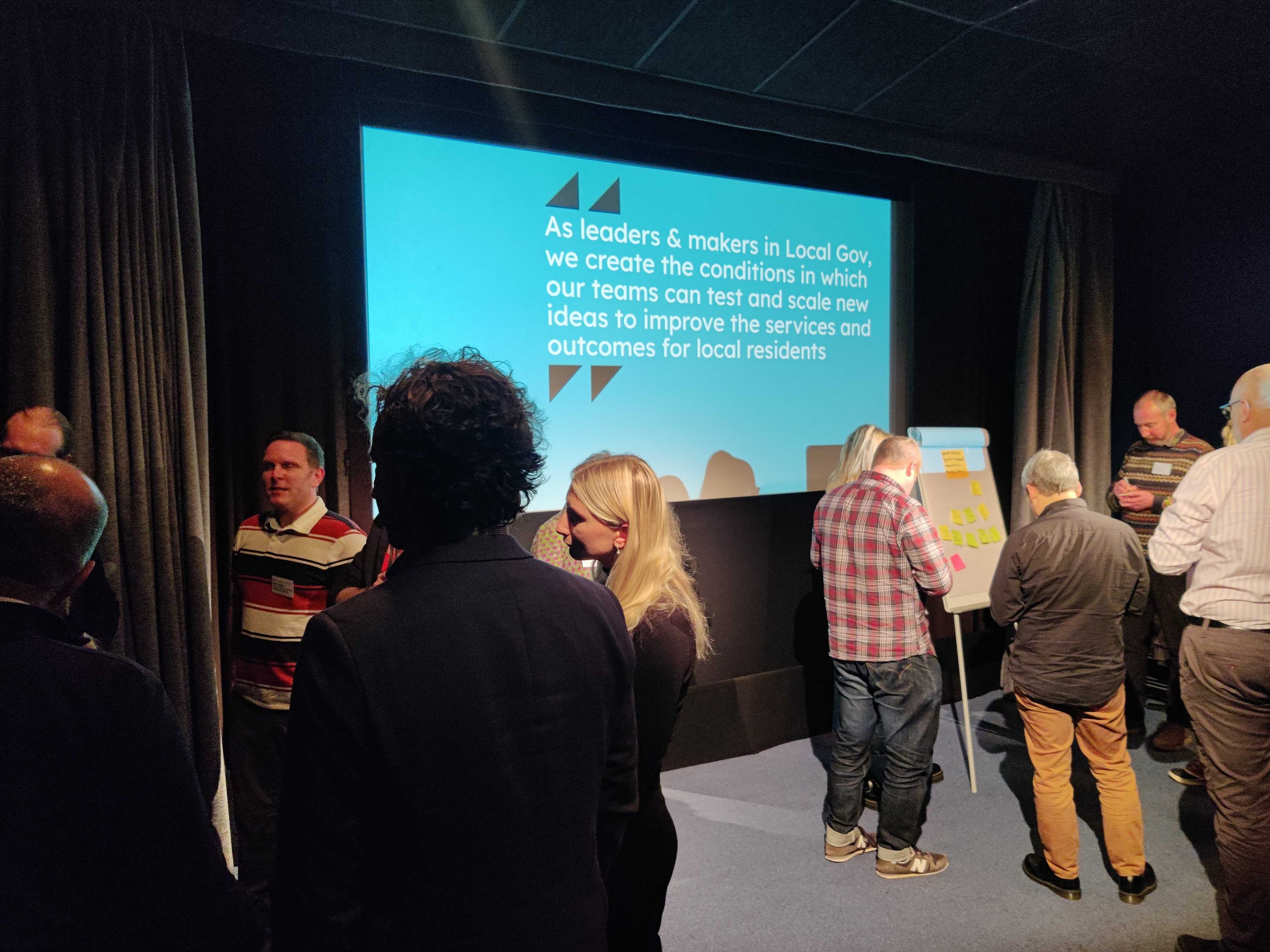 A few small groups of people mingling together alongside a big screen that reads "As leaders and makers in local government, we create the conditions for our teams to test and scale their ideas to deliver outcomes for local services"