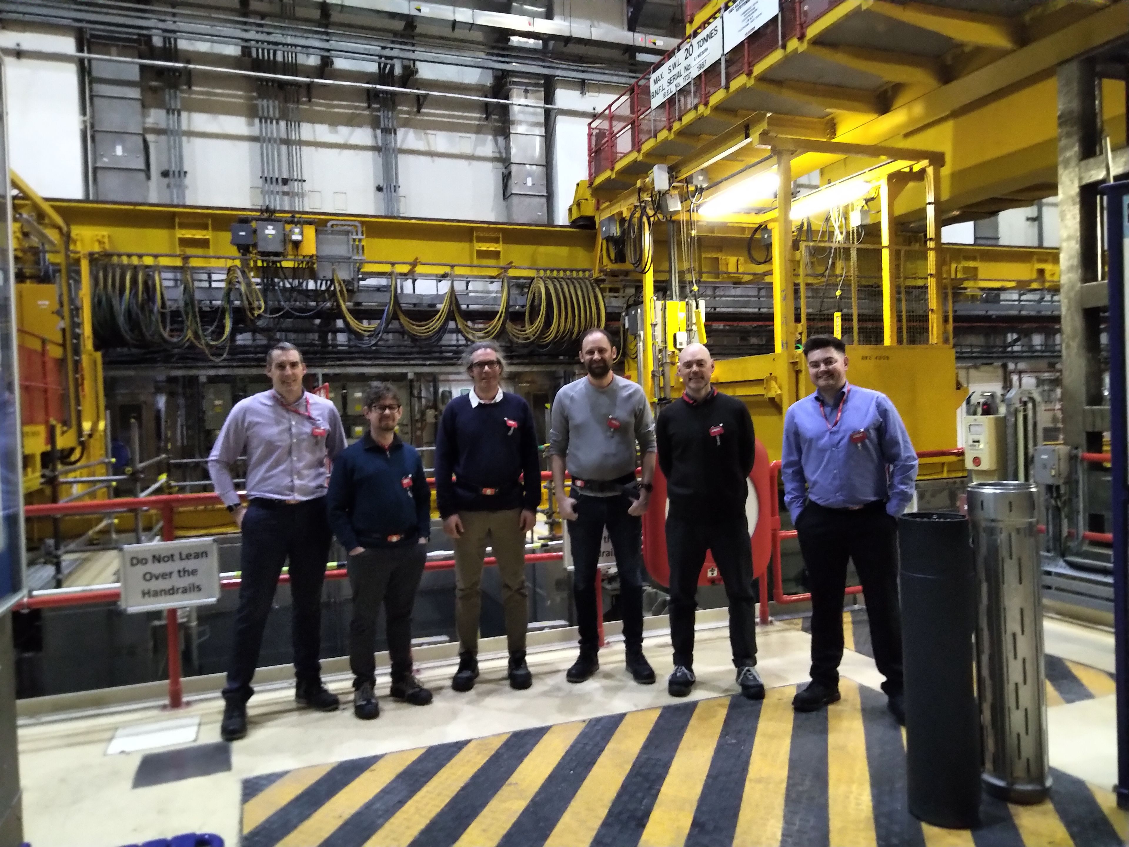 Six people stand to camera in the middle of a nuclear processing facility with a dominating yellow and black colour scheme