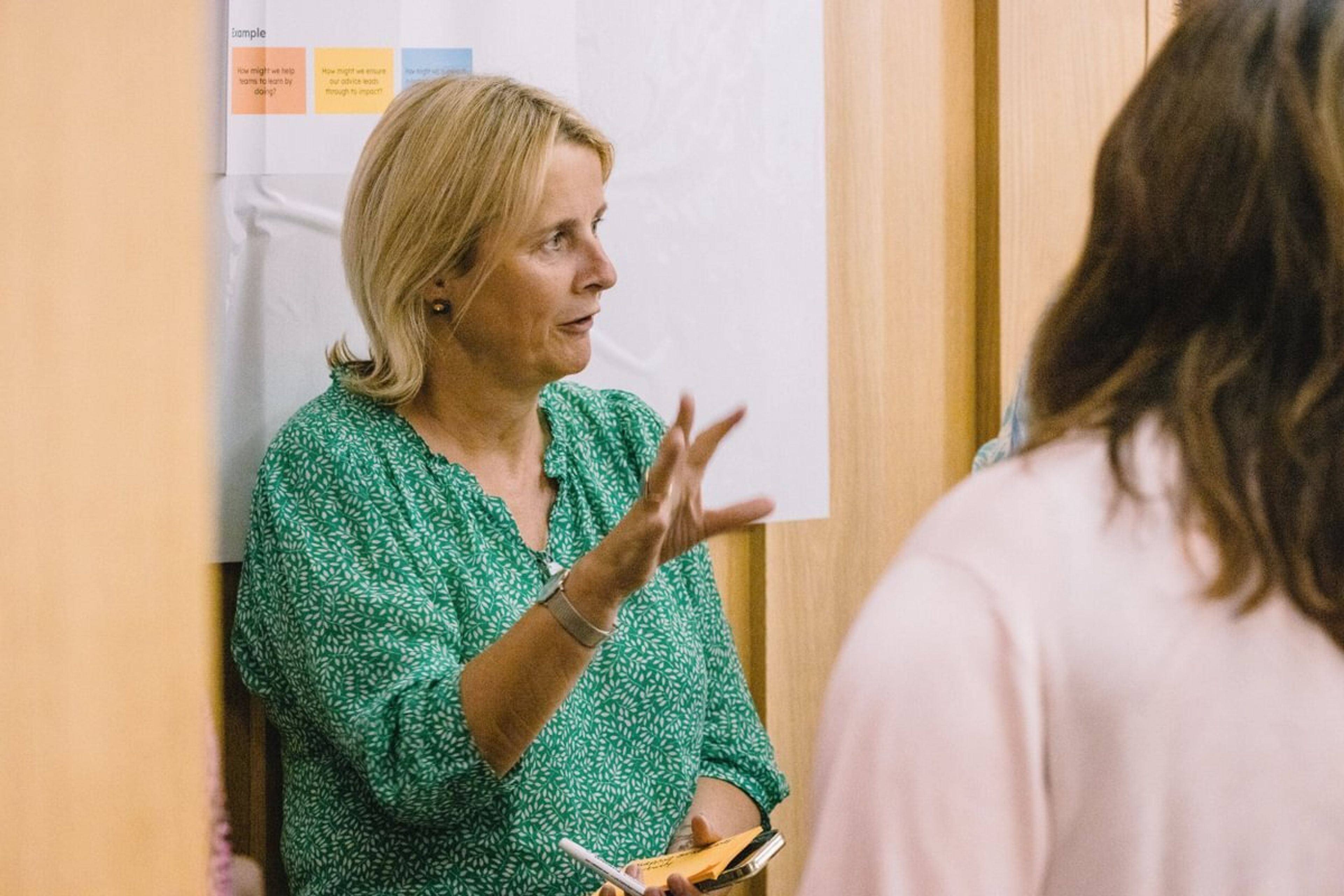 A woman is leaning against a wall while addressing a group of people, using hand gestures that suggest she is explaining something