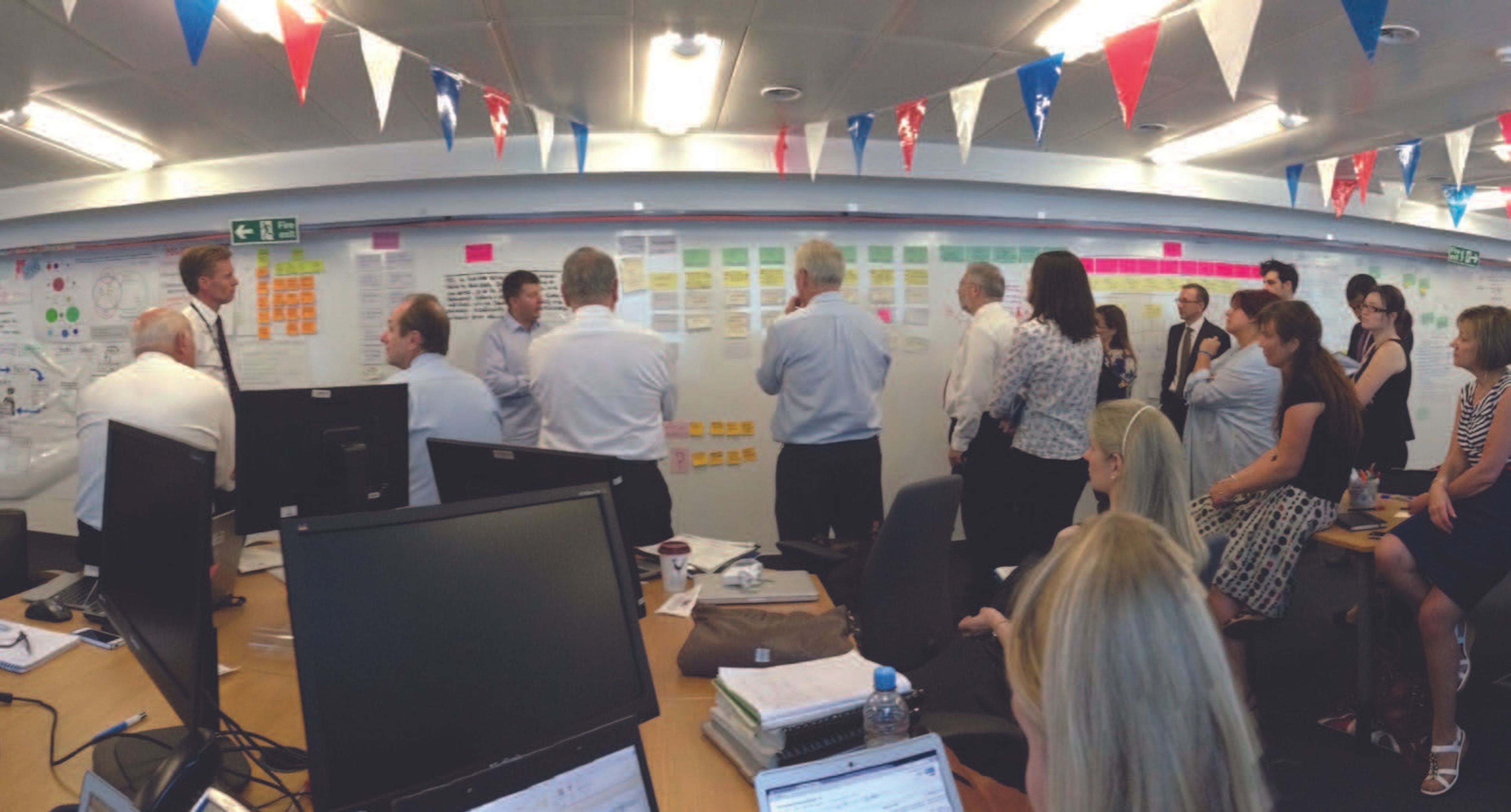 A room with over a dozen people huddle around a wall where someone is talking to the group, surrounded by paper artefacts and with bunting on the ceiling with the colours of the United Kingdom