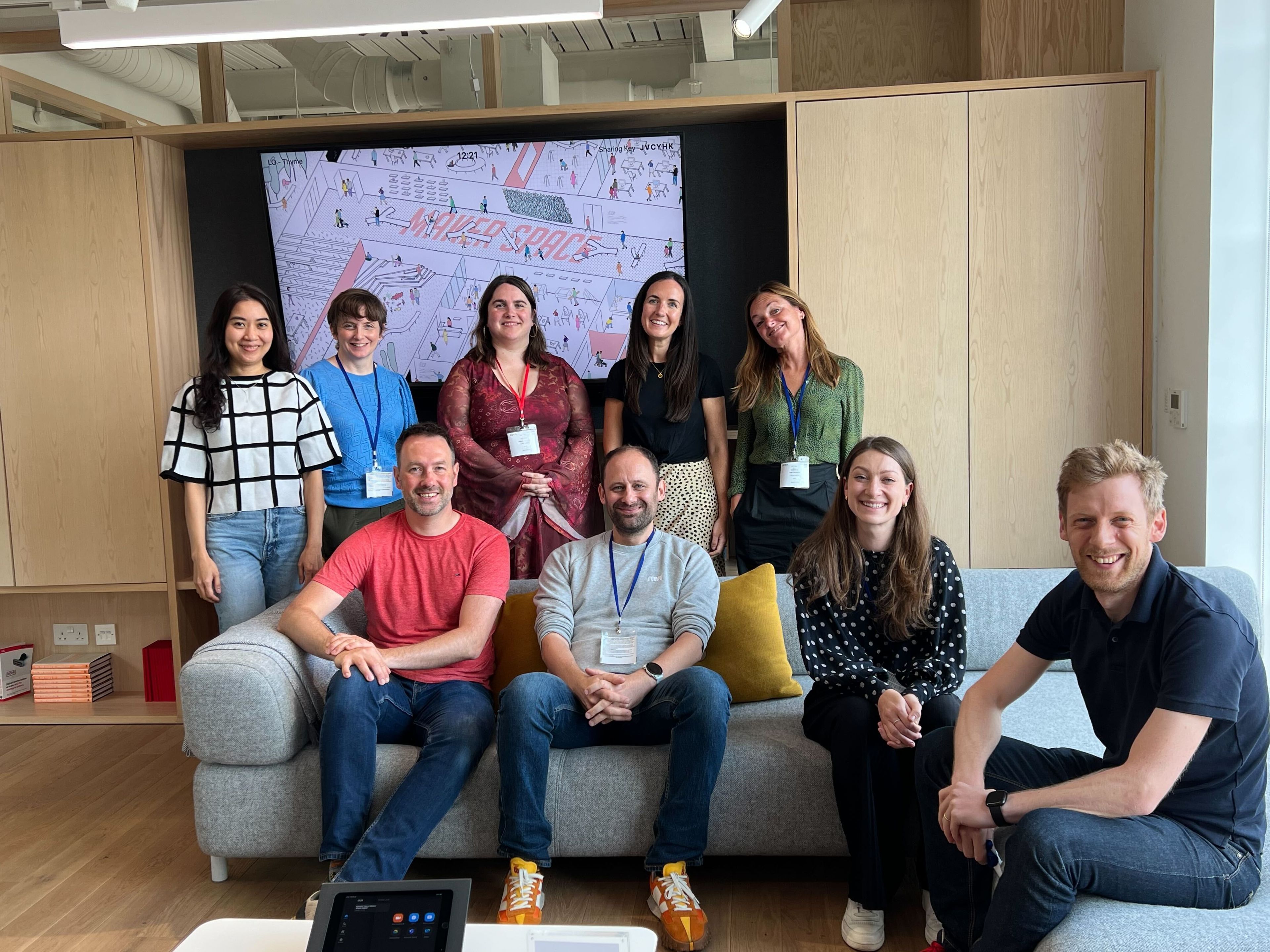 A team of consultants with some sitting in a sofa and some standing, all looking at camera and smiling in a relaxed stance