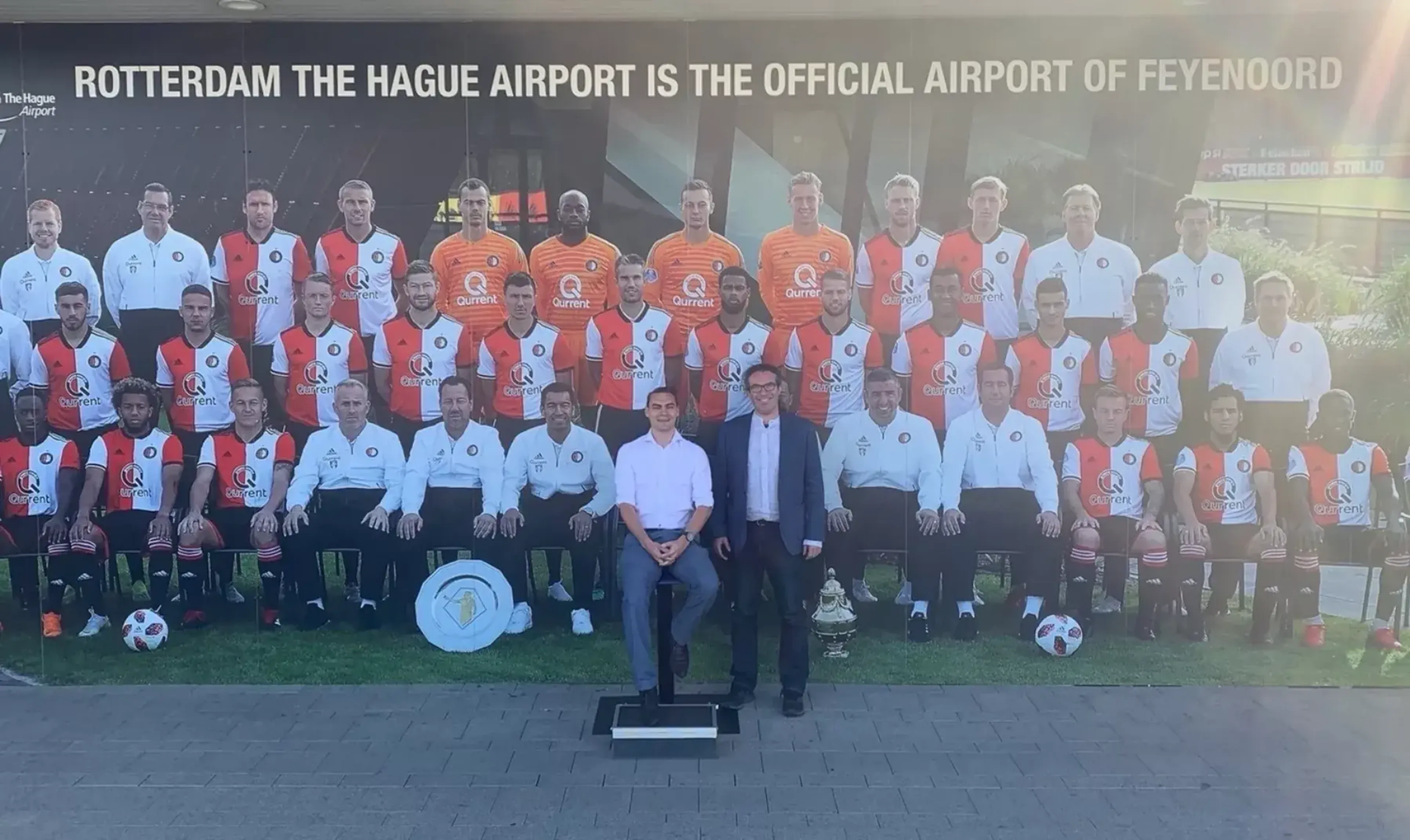 Andrew and James posing in front of a poste of Feyenoord football club in The Hague airport