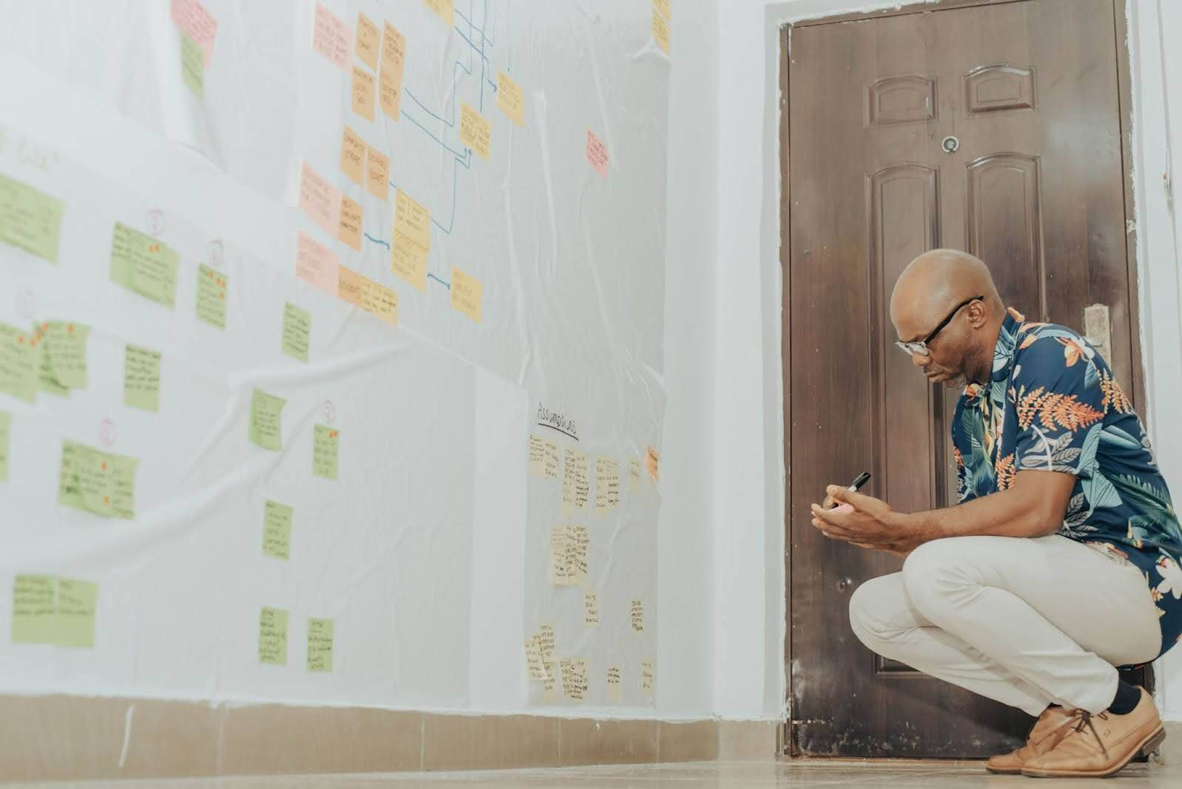 A person is crouched on the floor taking notes based on what the notes he's observing on the wall