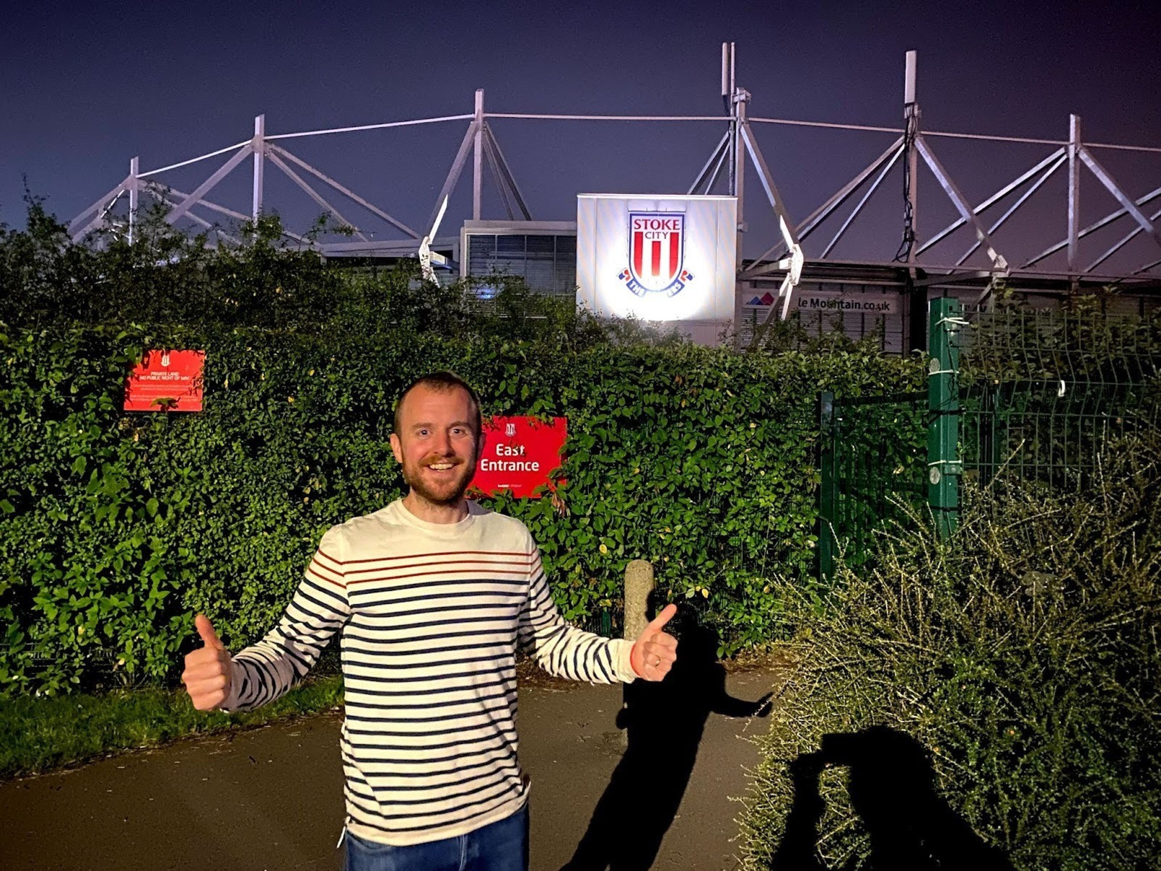 A man poses in front Stoke City F.C stadium with thumbs up and a wide smile