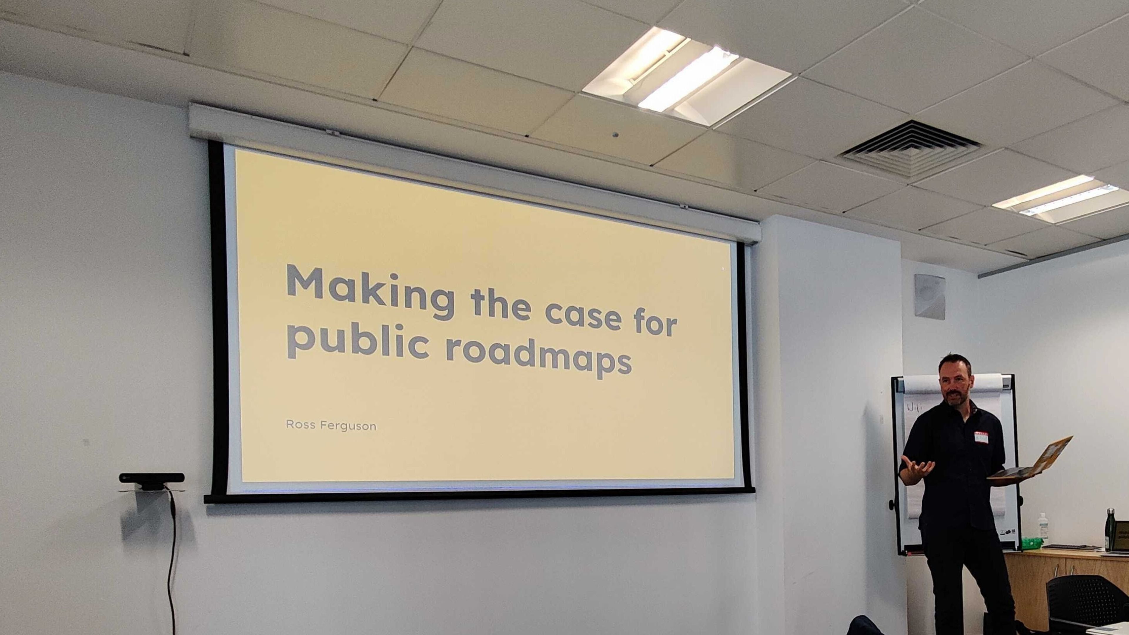 A presenter delivering a talk next to a big screen that reads "Making the case for public roadmaps"