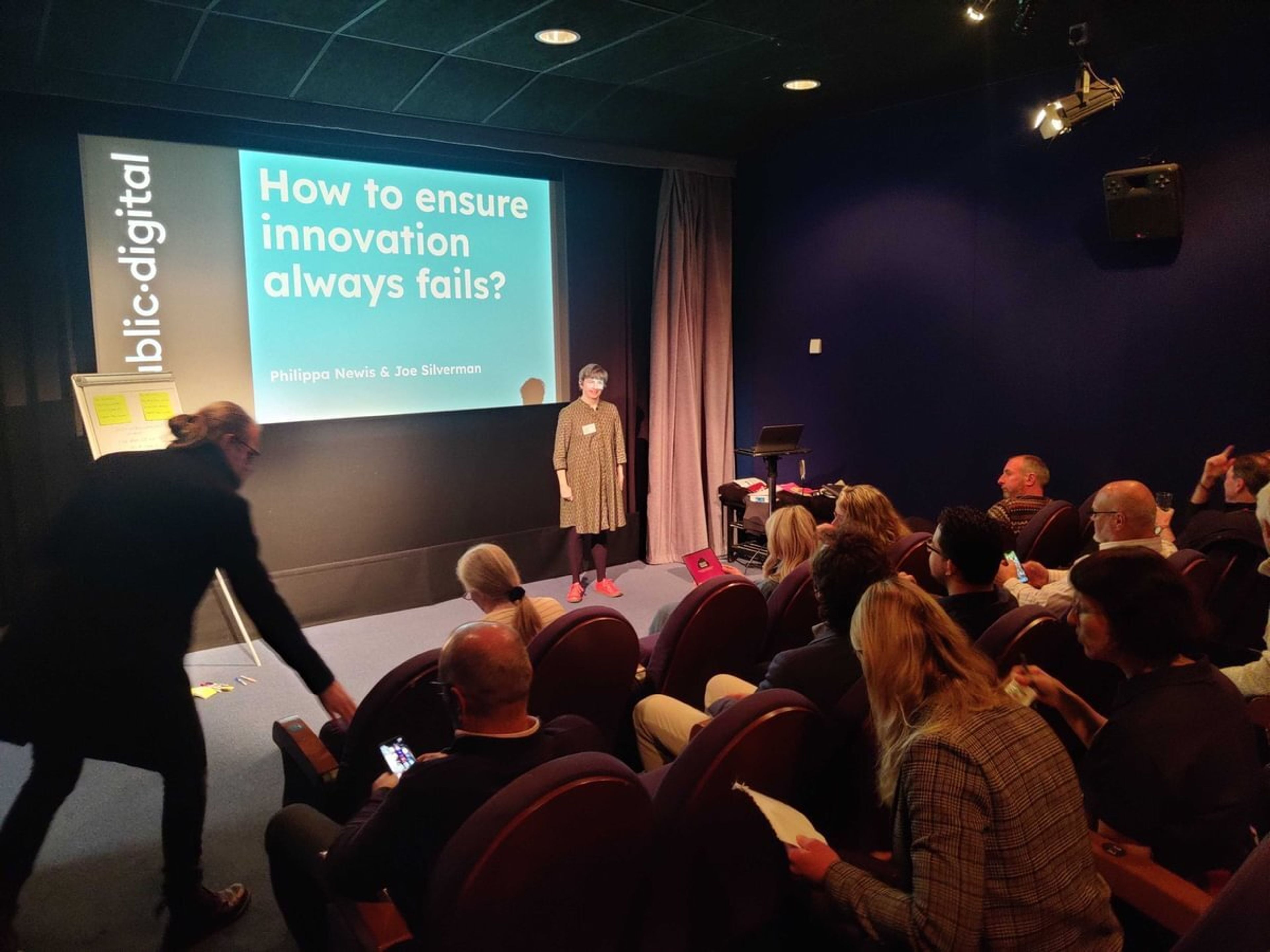 A woman presenting to an audience next to a screen that reads "how to ensure innovation always fails"