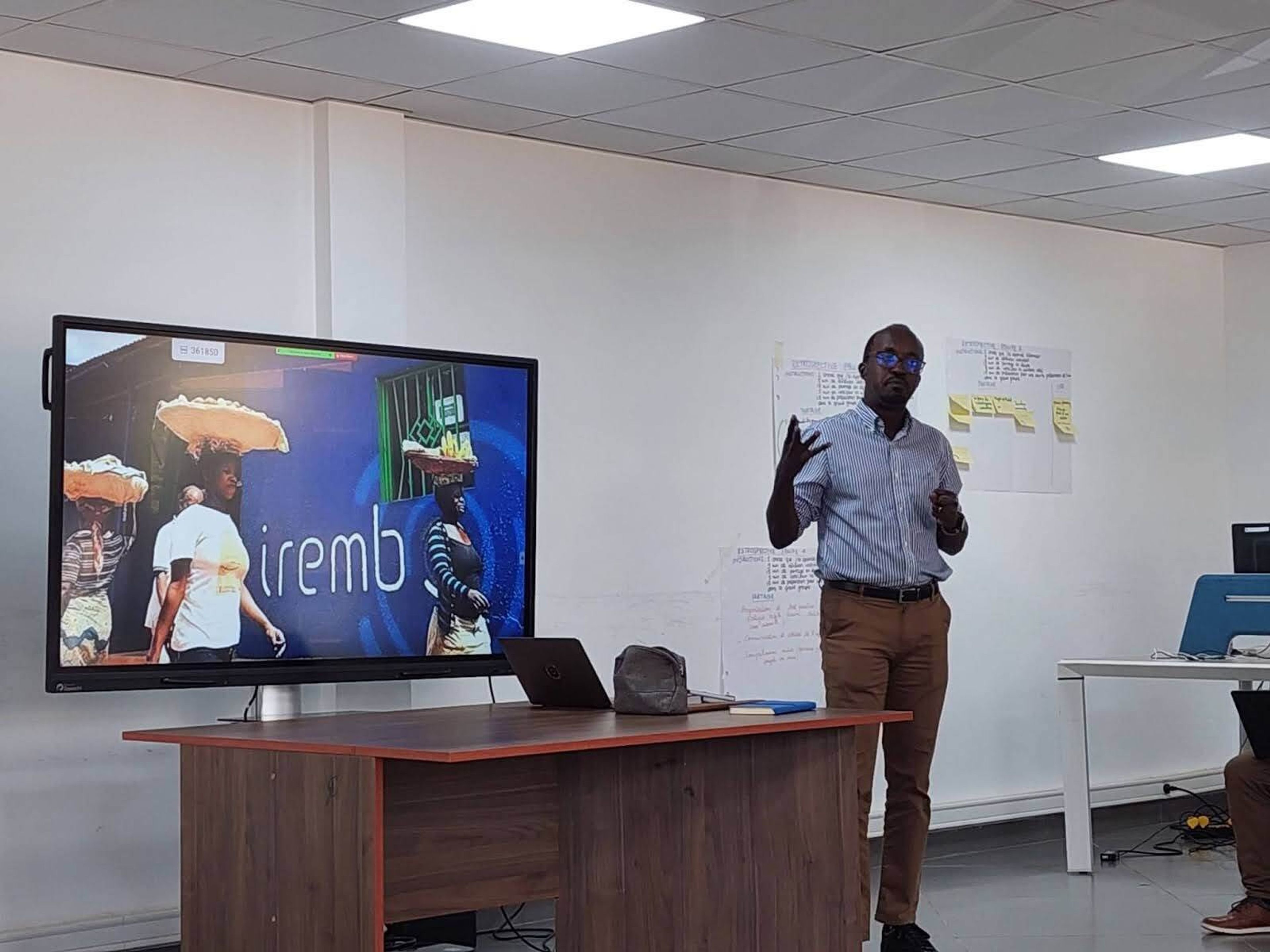 A man addresses an implied audience in an office room alongside a screen showing a logo for Irembo, government's online portal in Rwanda