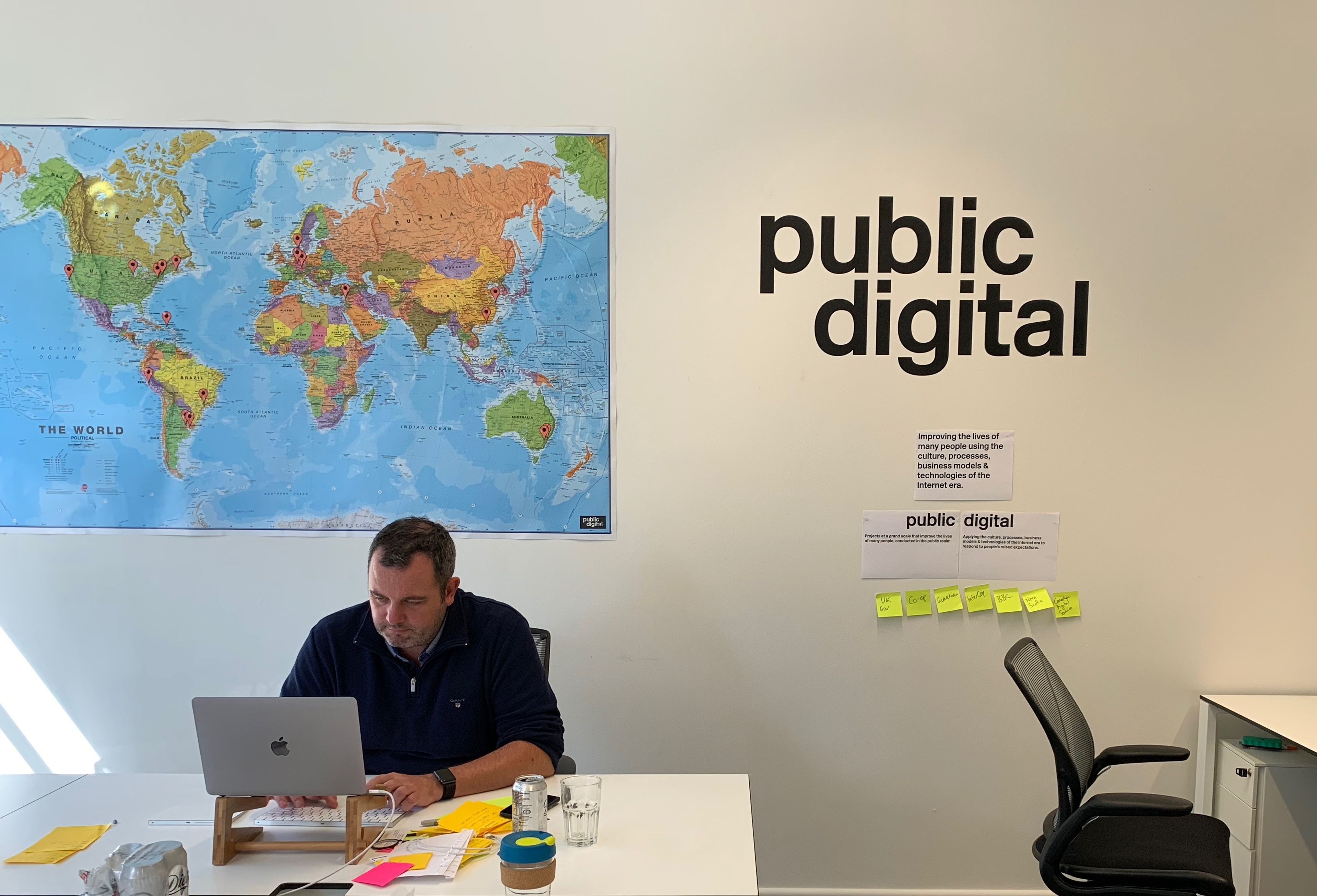 A man sitting on a desk with a laptop with a world map behind him and the Public Digital's logo